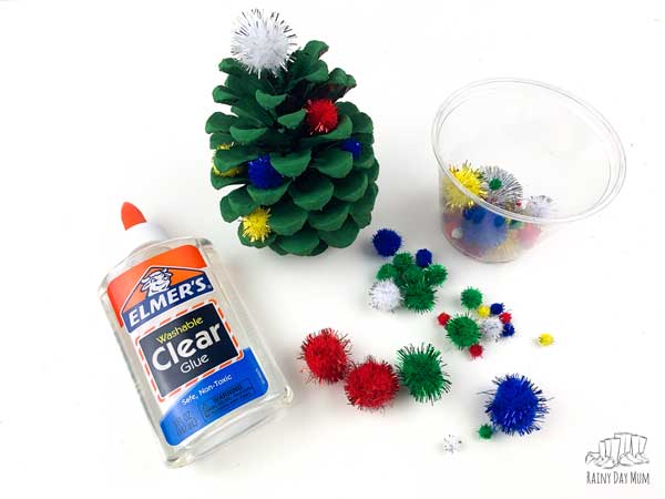 adding pom poms to a painted pine cone and gluing in place with Elmer Clear Glue