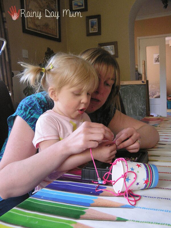 mum and toddler girl making a paper cup wind chime together at a table in the house