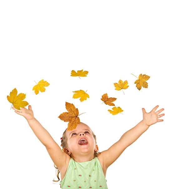 A preschool girl playing in fall leaves throwing in the air with a white background behind.