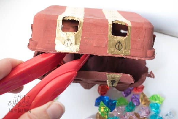 a pair of large kids tongs taking pirate gems and placing them in a handmade pirate treasure chest from an egg carton box