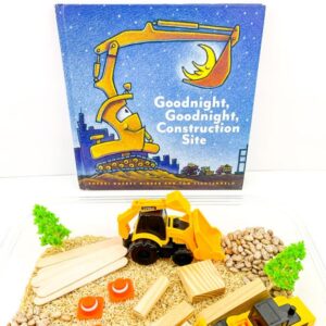 goodnight goodnight construction site board book placed above a simple construction themed sensory tub set up ready for a child to play. The sensory tub is filled with trucks, blocks, cones and trees on a bed of rice and pinto beans ready for play.