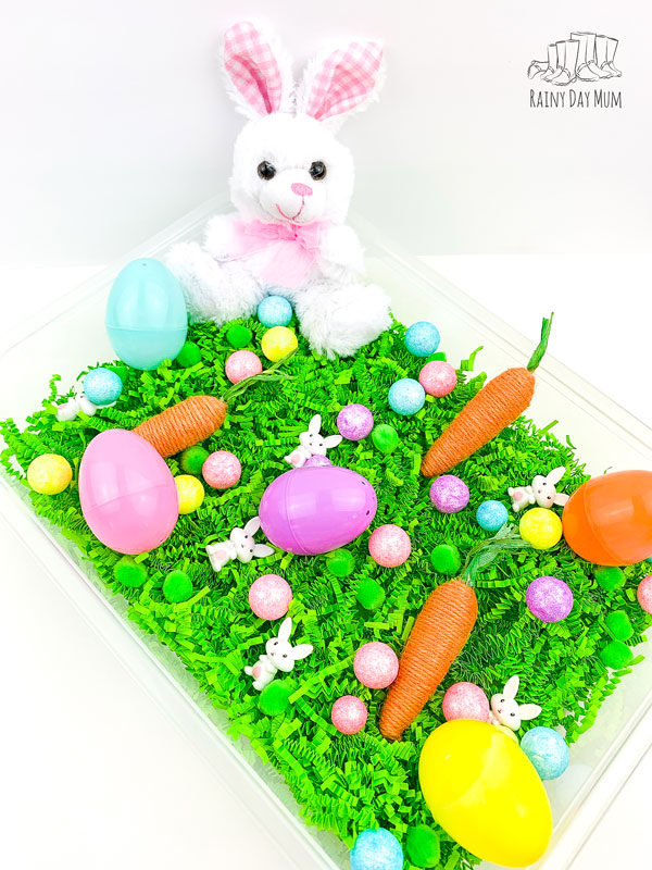 an invitation to play set up for a preschooler to play in a sensory tub, there is a plush easter bunny, plastic easter eggs, jute wrapped artificial carrots, glitter balls and mini bunnies on top of Easter grass 
