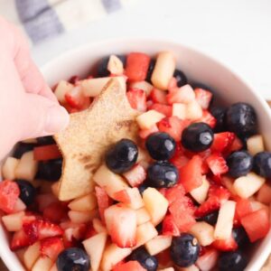 a hand dipping a homemade star shaped cinnamon coated tortilla chip into a bowl with a red white and blue fruit salsa in , you can see blueberries strawberries, watermelon and apple slices in the bowl