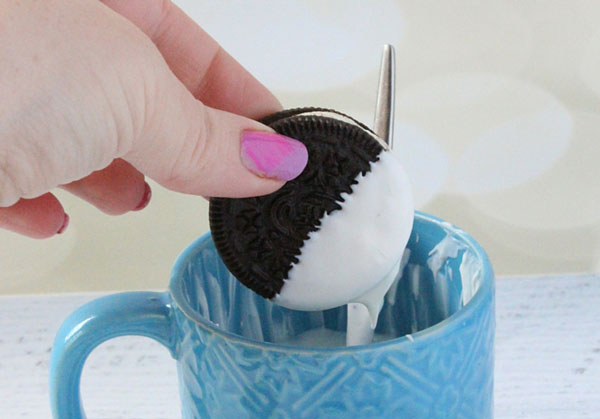 a female hand holding an Oreo which has been dipped into the mug below it full of white-candy melt, the Oreo is half covered and some is dripping off