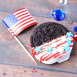 image of an Oreo Cookie which has been dipped in white chocolate and drizzled with red candy with star sprinkles added as a 4th of July Treat for kids to make next to a US flag on a wooden table top a few blue, red and white gems are scattered around