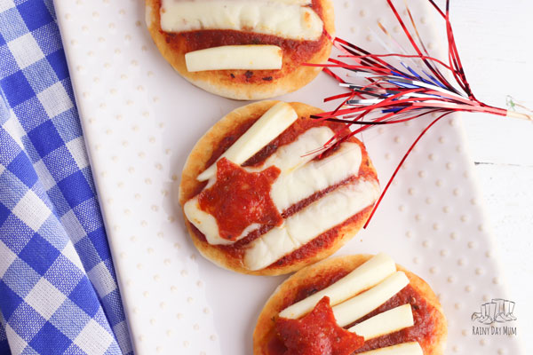 3 mini flag pizzas with cheese string stripes and mini pepperoni star on a white plate with a sparkler laying on a blue gingham cloth