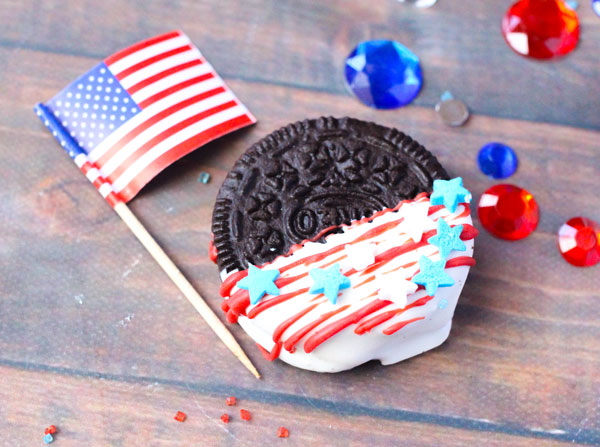 simple Oreo Cookie half dipped in white Candy melts with drizzled stripes of red across and blue and white stars sprinkled on top, beside it are red and blue gems and a US flag